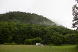 Experience nature with an RV rental! White RV & car parked in a lush green field before a misty, forested mountain.