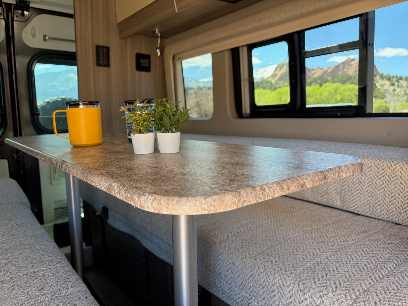 RV rental interior: a dining table with a mug and plants, flanked by comfortable benches and scenic windows with mountain views.