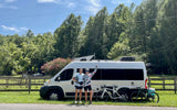Two women and bikes next to an RV rental, set for an outdoor cycling adventure. Sunny day, trees in back.