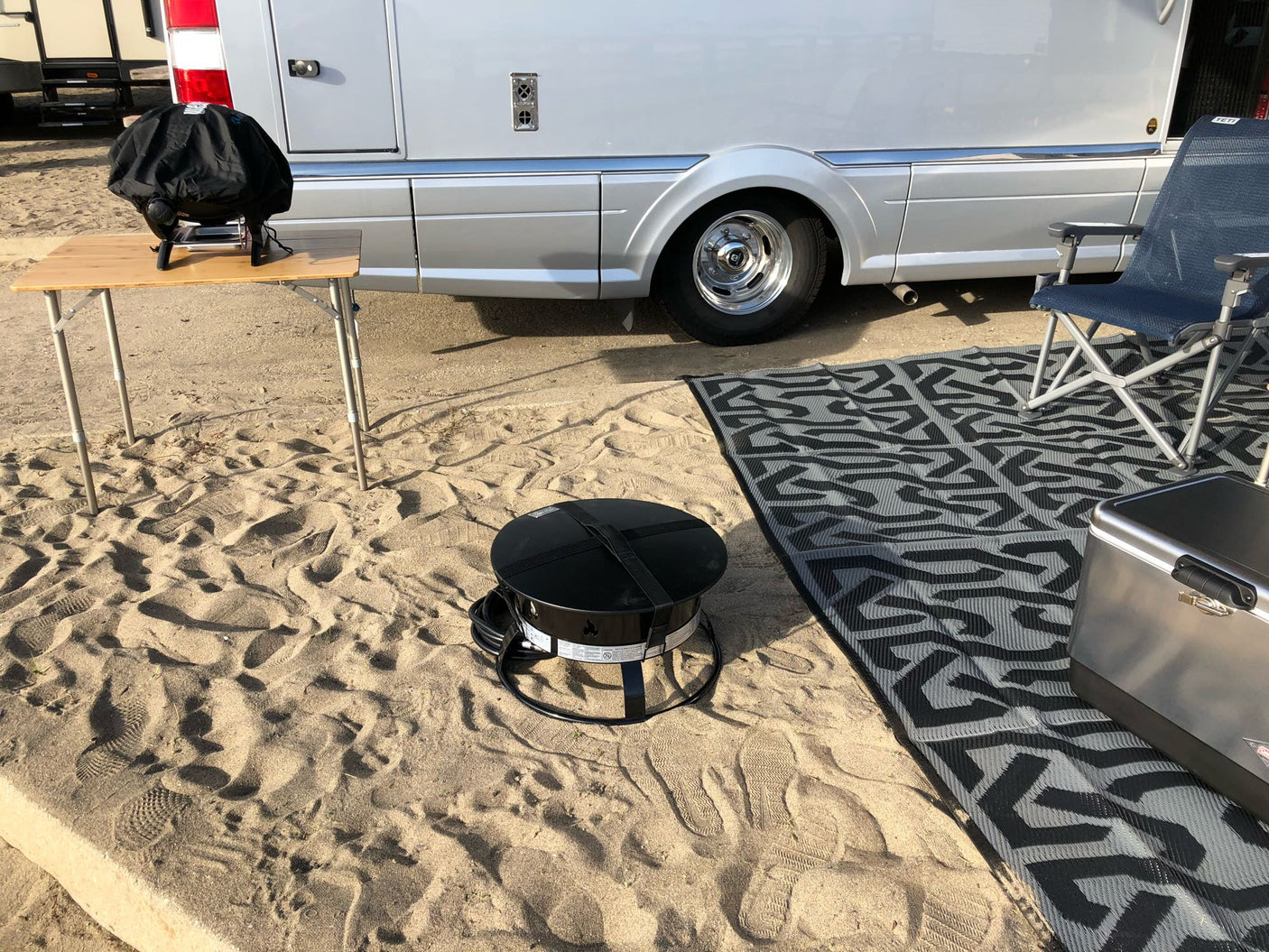 RV rental on sand: motorhome, covered grill on table, portable fire pit, mat, chair, & cooler for outdoor living.