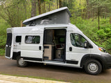 White Winnebago Solis RV rental camper van with pop-top open, parked in a lush green forest. Interior visible.