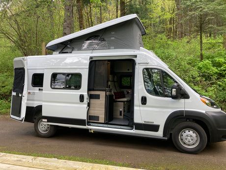 White Winnebago Solis RV rental camper van with pop-top open, parked in a lush green forest. Interior visible.