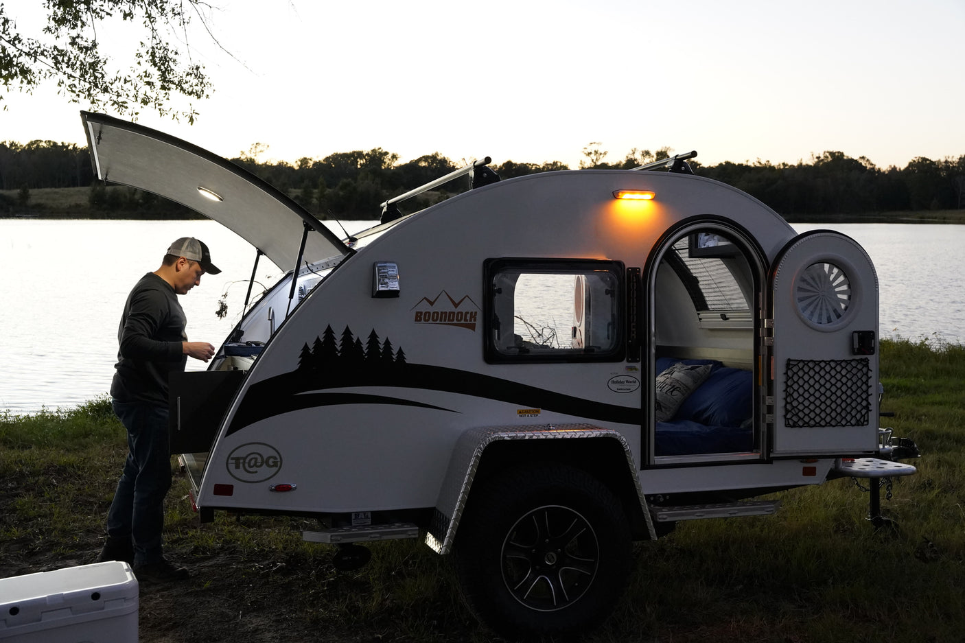 A man utilizes the outdoor galley of his teardrop camper RV rental by a lake, ideal for serene outdoor adventures.