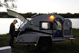 A man utilizes the outdoor galley of his teardrop camper RV rental by a lake, ideal for serene outdoor adventures.