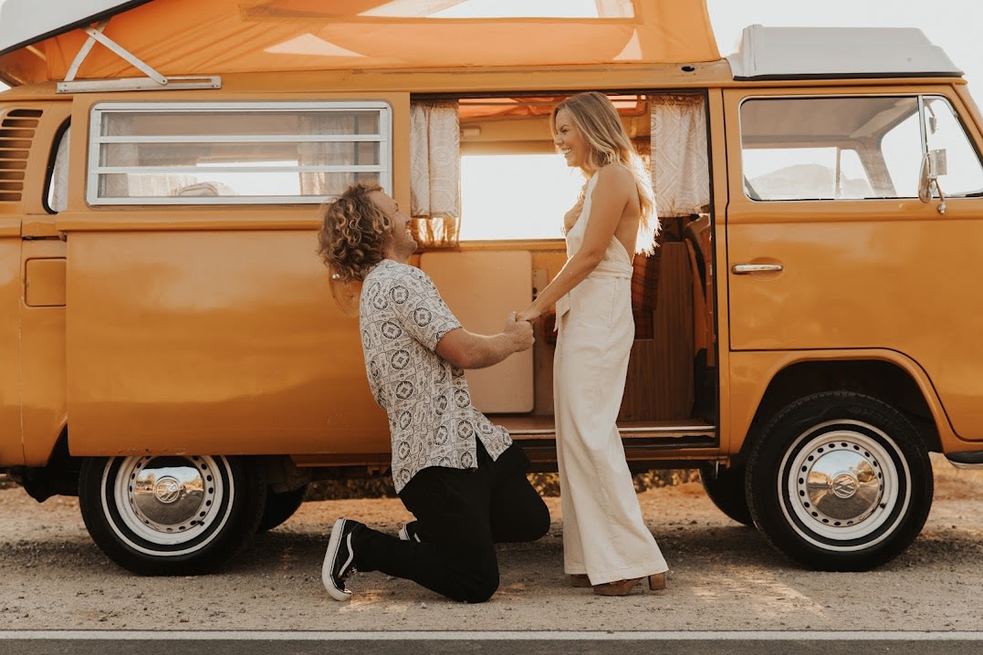 Romantic proposal by an orange RV rental. Man on knee holds woman's hands beside a classic camper van at sunset.