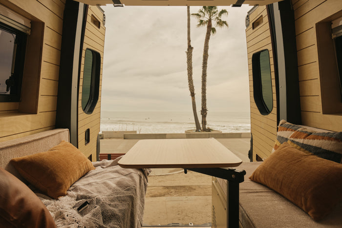 Cozy RV rental interior with open back doors revealing an ocean view, palm trees, and surfers. Features a table, cushions, & blanket.