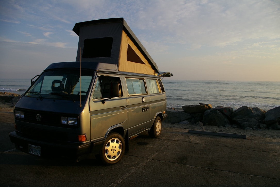  enjoying sunset over ocean waves. Perfect for road trip adventures.|Pop-top VW camper van parked on pavement by the ocean. A perfect RV rental for scenic beachfront trips.|Front of a blue-grey VW van — photo 5