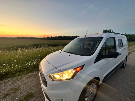 White camper van by wildflower field at sunset, headlights on. Great for an RV rental escape.