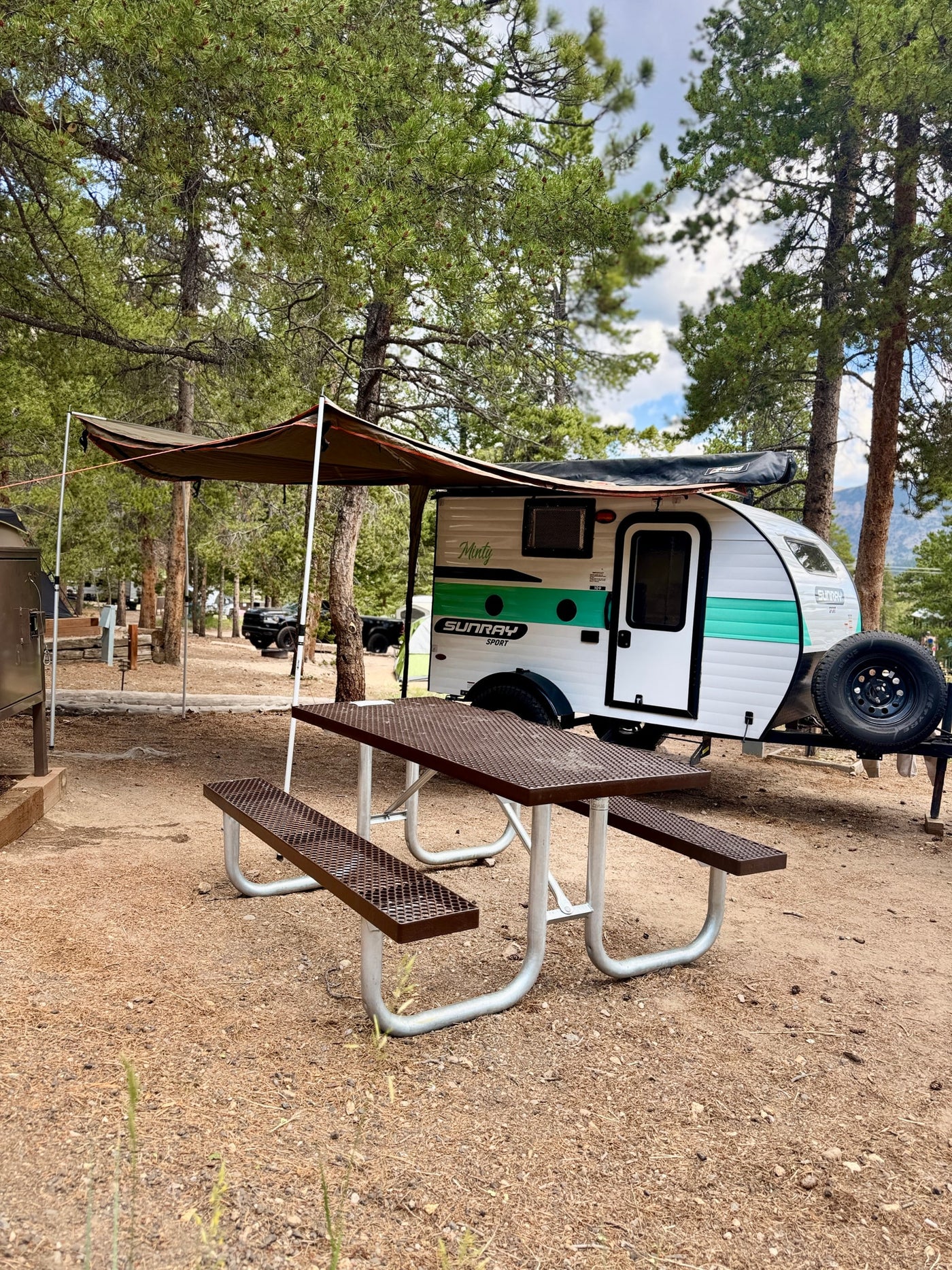 A "Minty" Sunray Sport teardrop RV rental with awning and picnic table, nestled among pine trees at a cozy campsite. — photo 4