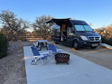 RV rental campsite setup: A black camper van with an awning, picnic table, chairs, and fire pit on a concrete pad.