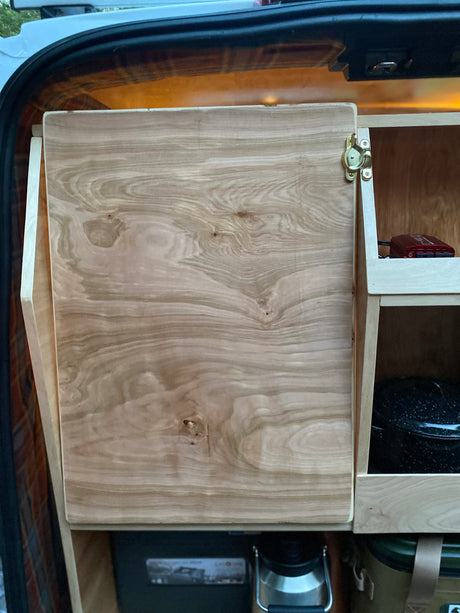 RV rental custom cabinetry: a light wood door with a brass latch opens to reveal interior storage, with open shelves holding essentials.
