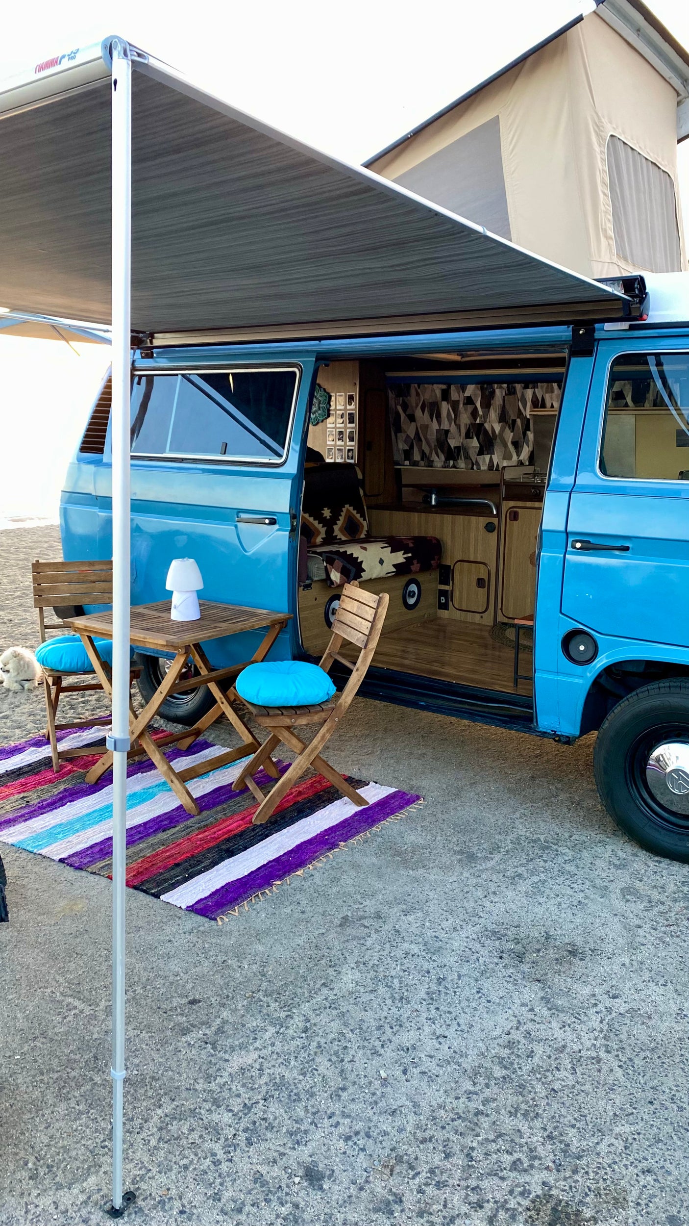 This blue camper van with an extended awning offers a cozy outdoor living space with table, chairs, and colorful rug, ideal for an RV rental adventure.