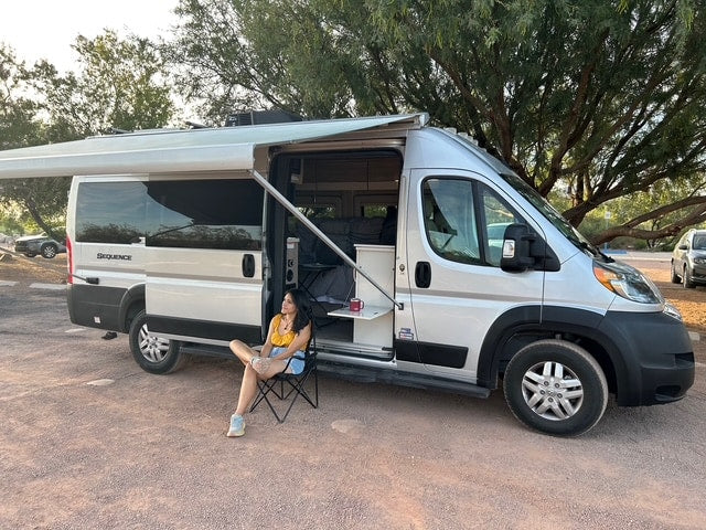 Enjoy a relaxing outdoor moment with an RV rental. A woman sits by a silver camper van with its awning extended in a treed area.
