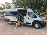 Enjoy a relaxing outdoor moment with an RV rental. A woman sits by a silver camper van with its awning extended in a treed area.