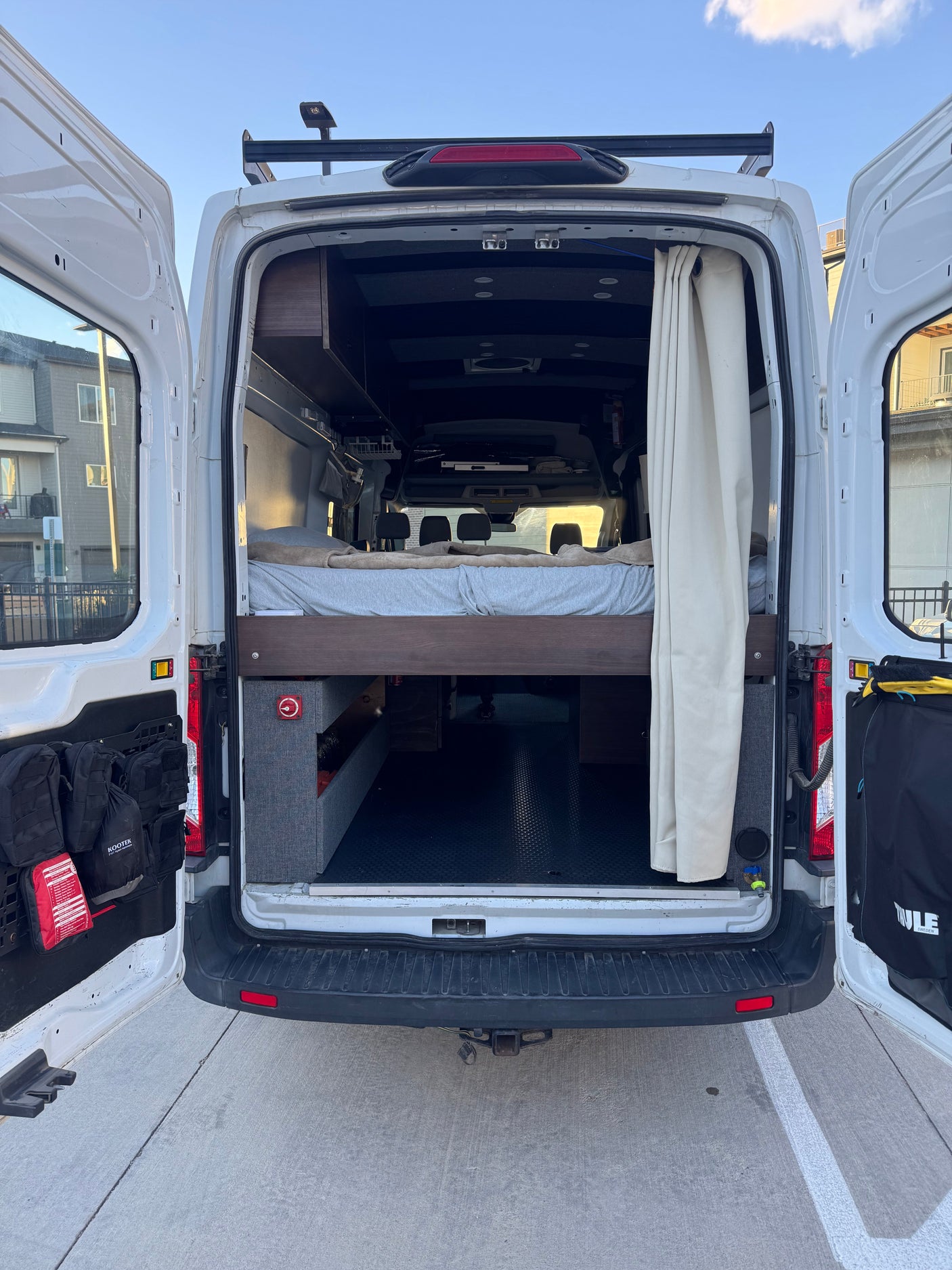 Open rear doors of a white camper van reveal a custom interior with a bed, storage, and curtain. Perfect for an rv rental adventure.