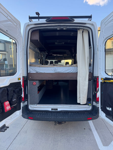 Open rear doors of a white camper van reveal a custom interior with a bed, storage, and curtain. Perfect for an rv rental adventure.