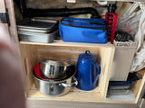 RV rental kitchen: Compact storage with pots, pans, a HotLogic bag, French press, and various bakeware in a cupboard.