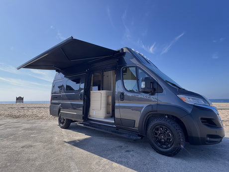 Gray camper van (RV rental) with open side door & extended awning, parked by a sunny beach and ocean. Ready for adventure!