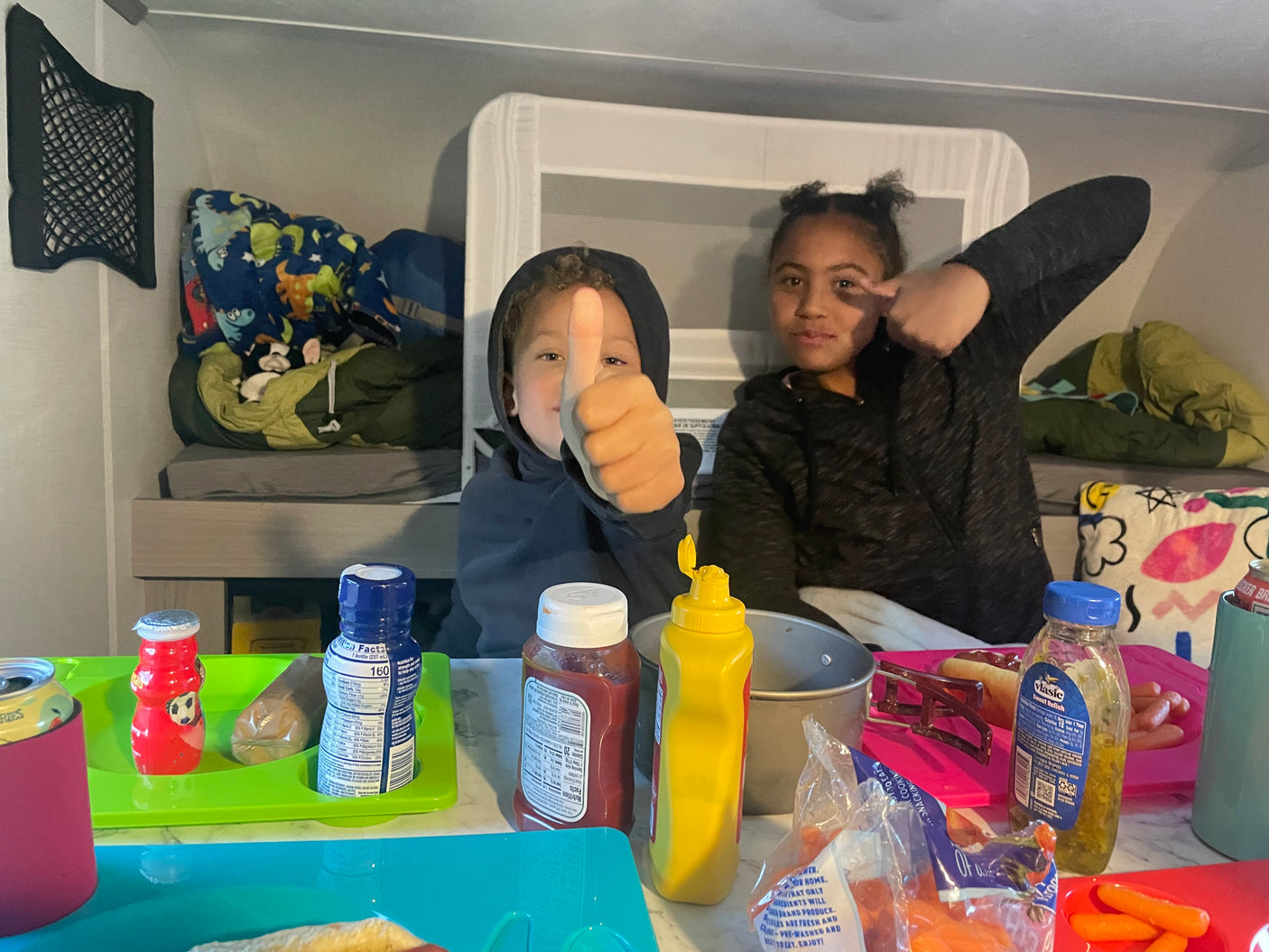 Inside an RV rental: two happy kids enjoy a hot dog meal in the bunk area, perfect for family adventures.