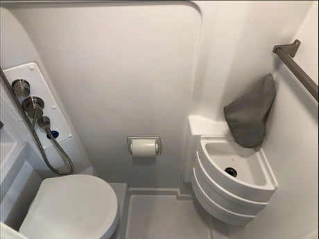 View of a compact RV rental wet bath with toilet, wall-mounted shower controls, corner sink, and toilet paper holder.