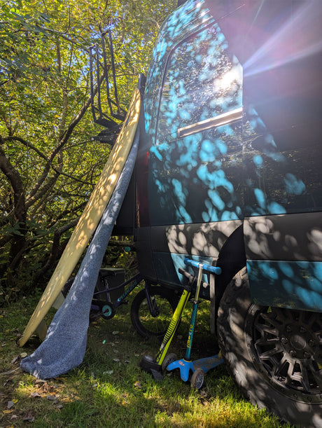 RV rental parked in dappled sunlight with a surfboard, kids' bike, and scooters by green trees.