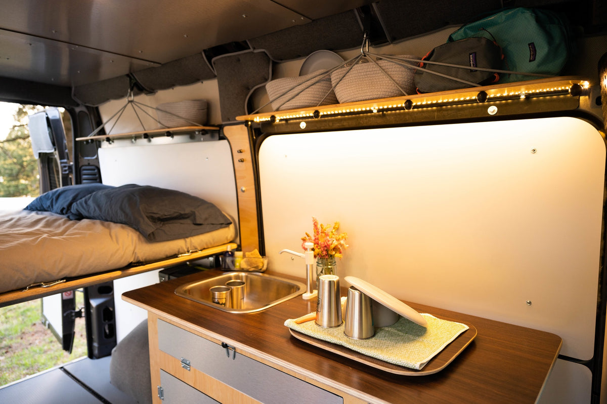 Cozy rv rental interior, ideal for van life, with a kitchen counter, sink, bed, and illuminated overhead storage. — photo 9
