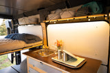 Cozy rv rental interior, ideal for van life, with a kitchen counter, sink, bed, and illuminated overhead storage.
