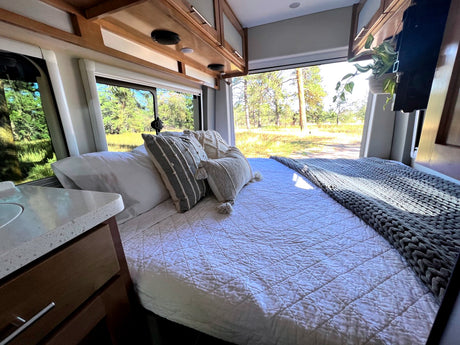 Cozy RV rental interior: comfortable bed with white bedding, decorative pillows & knit blanket, offering serene forest views outside.
