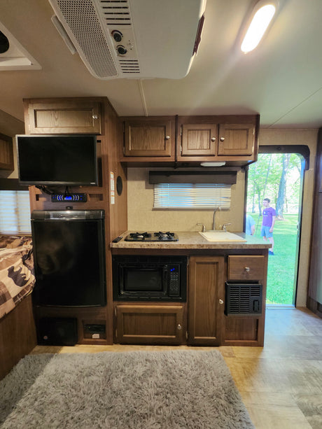 RV rental kitchen area with TV, fridge, and microwave. An open door reveals a sunny outdoor scene with people.