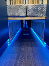 Modern RV rental interior: wood-look floor, comfortable bed, and striking blue LED ambient lighting line the aisle.