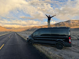 A person with outstretched arms on a black RV rental van, celebrating a scenic mountain and cloud vista on a remote road.