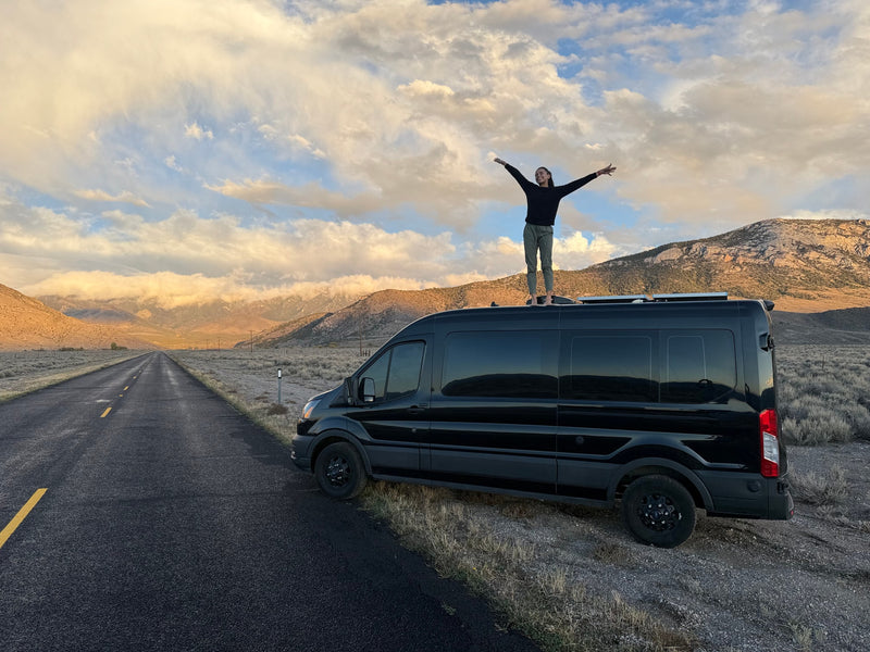 A person with outstretched arms on a black RV rental van, celebrating a scenic mountain and cloud vista on a remote road. — photo 9