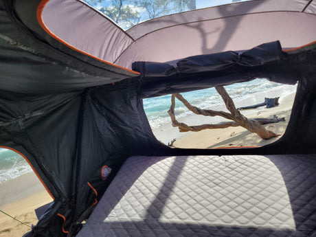 View from an RV rental's rooftop tent on a sandy beach, showcasing ocean waves and driftwood. Enjoy seaside camping comfort.