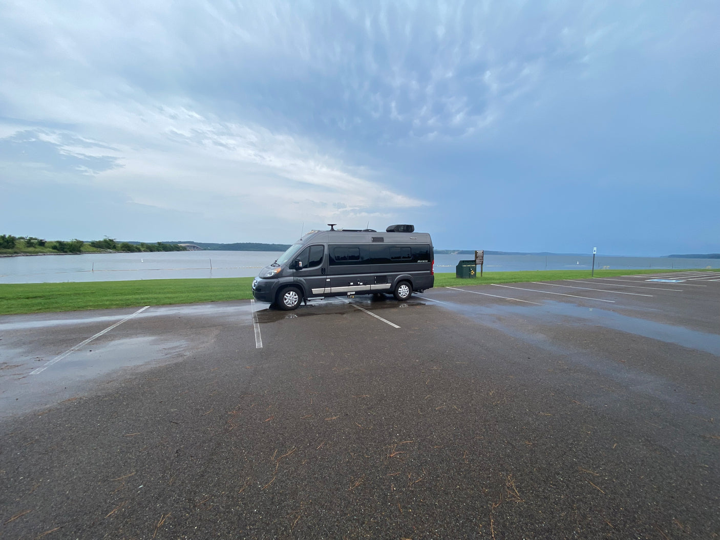 Gray rv rental camper van parked on a wet asphalt lot next to a lake under cloudy skies, ready for adventure.