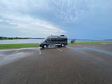 Gray rv rental camper van parked on a wet asphalt lot next to a lake under cloudy skies, ready for adventure.