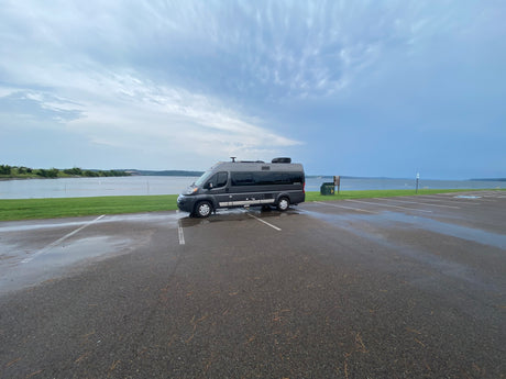 Gray rv rental camper van parked on a wet asphalt lot next to a lake under cloudy skies, ready for adventure.
