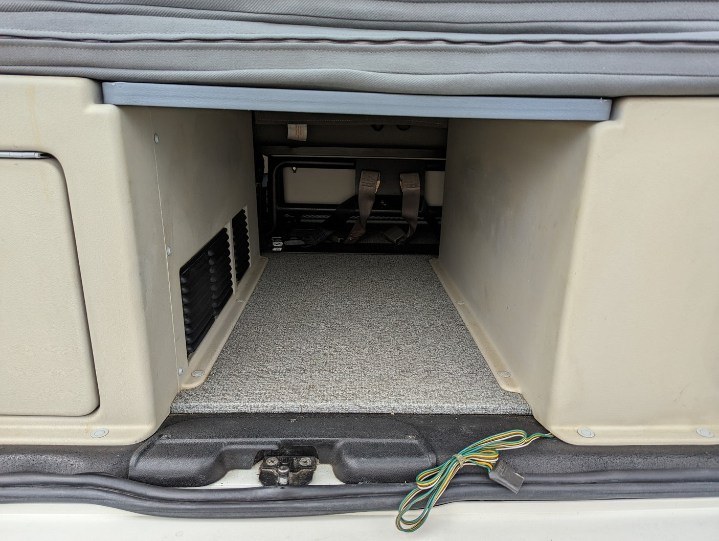 RV rental: Rear cargo area under a bench, featuring a carpeted floor, beige walls with vents, and trailer wiring.