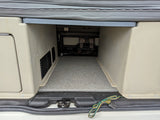 RV rental: Rear cargo area under a bench, featuring a carpeted floor, beige walls with vents, and trailer wiring.