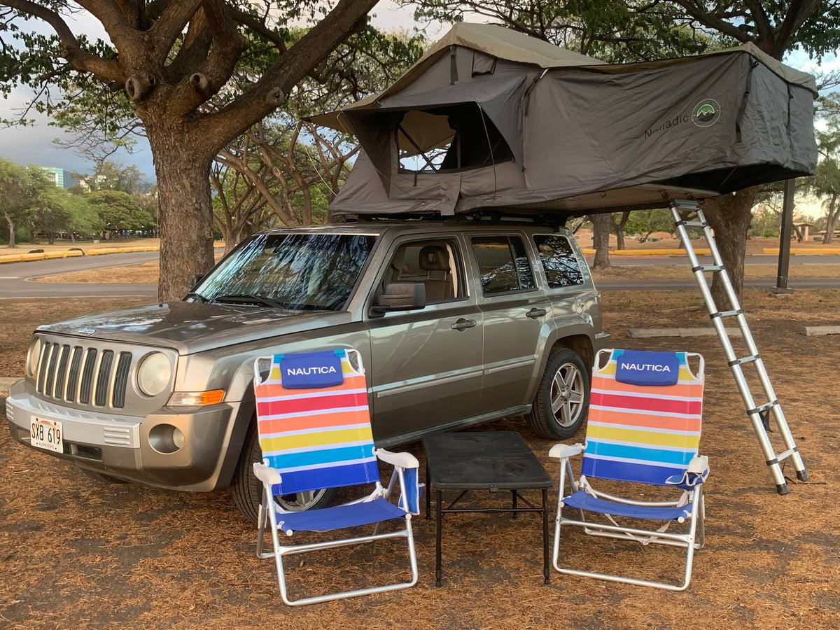 A grey Jeep Patriot with a deployed 'Nomadic' rooftop tent & ladder, plus colorful Nautica chairs. A great RV rental option for camping. — photo 8