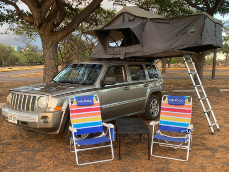 A grey Jeep Patriot with a deployed 'Nomadic' rooftop tent & ladder, plus colorful Nautica chairs. A great RV rental option for camping.