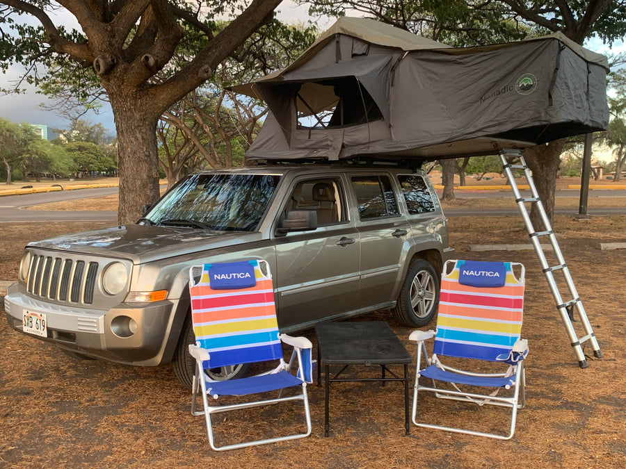 A grey Jeep Patriot with a deployed 'Nomadic' rooftop tent & ladder, plus colorful Nautica chairs. A great RV rental option for camping.
