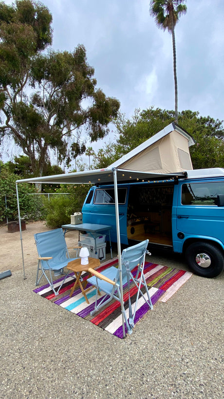 Blue camper van RV rental with pop-top, awning, chairs, table, and colorful rug set up outdoors under trees.