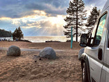 RV rental parked on a sandy beach at sunset, with sun rays over the lake and distant mountains. People relax near the water.