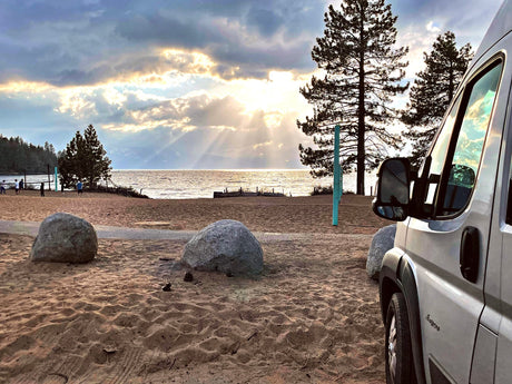 RV rental parked on a sandy beach at sunset, with sun rays over the lake and distant mountains. People relax near the water.