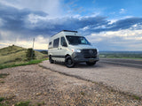 A light-colored Sprinter van, ideal for an RV rental, parked on a gravel shoulder with a scenic mountain view under dramatic clouds.