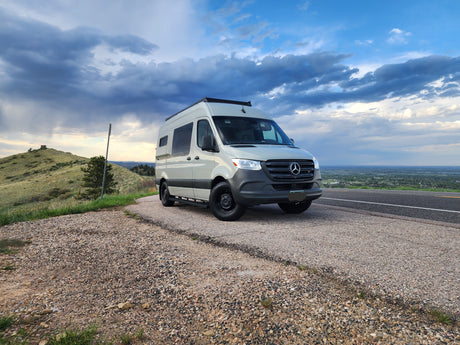 A light-colored Sprinter van, ideal for an RV rental, parked on a gravel shoulder with a scenic mountain view under dramatic clouds.