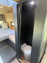 RV rental van conversion interior: Bed & private dark gray bathroom with composting toilet. Wood walls and ceiling.