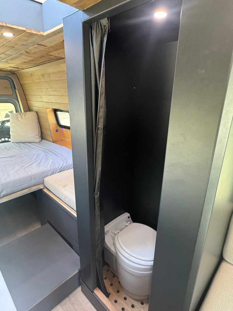RV rental van conversion interior: Bed & private dark gray bathroom with composting toilet. Wood walls and ceiling. — photo 18