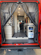 Spacious RV rental interior from the rear of a red camper van, showing a modular bed, kitchenette, and utility panel through mesh screens.