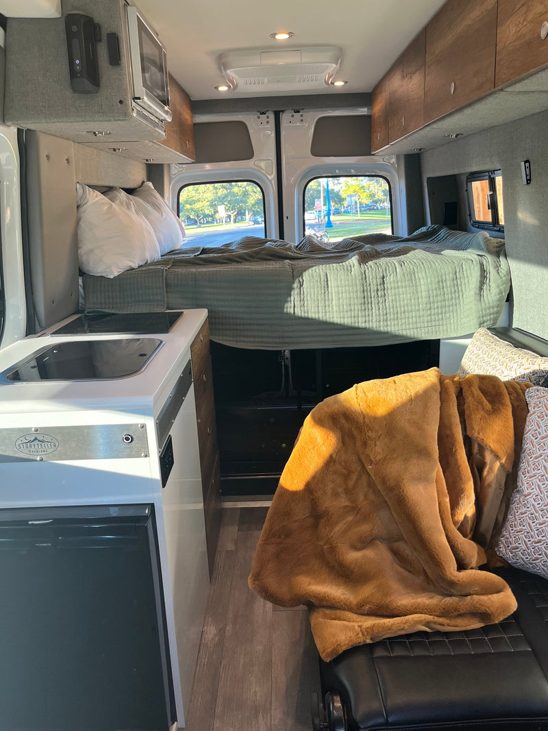 Bright rv rental camper van interior. Elevated bed with green quilt, functional kitchenette with sink, and comfy seating with blanket. — photo 33